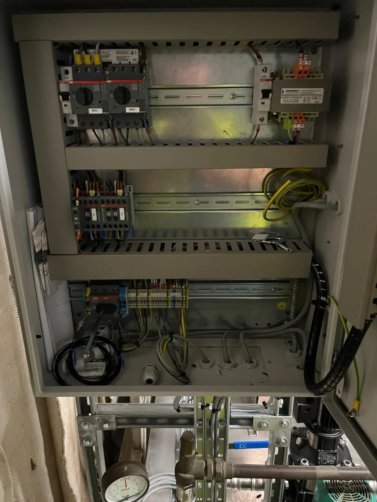 Open industrial control panel with motor starters, contactors, terminals and wiring on DIN rails, above plant-room pipework