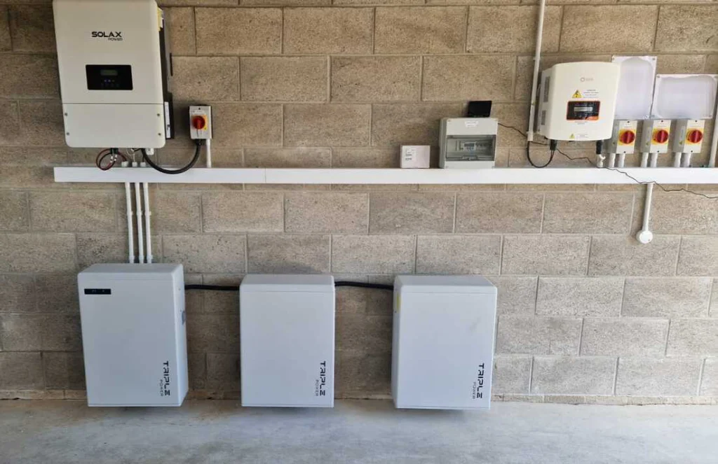 SolaX hybrid inverter and three wall-mounted SolaX Triple Power batteries on a block wall, with isolators and a generation meter